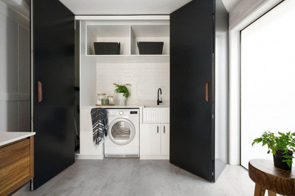 Contemporary single-wall laundry cupboard in Sydney with a farmhouse sink, flat-panel cabinets, white cabinets, white splashback, subway tile splashback, a side-by-side washer and dryer, grey floor and white benchtop.