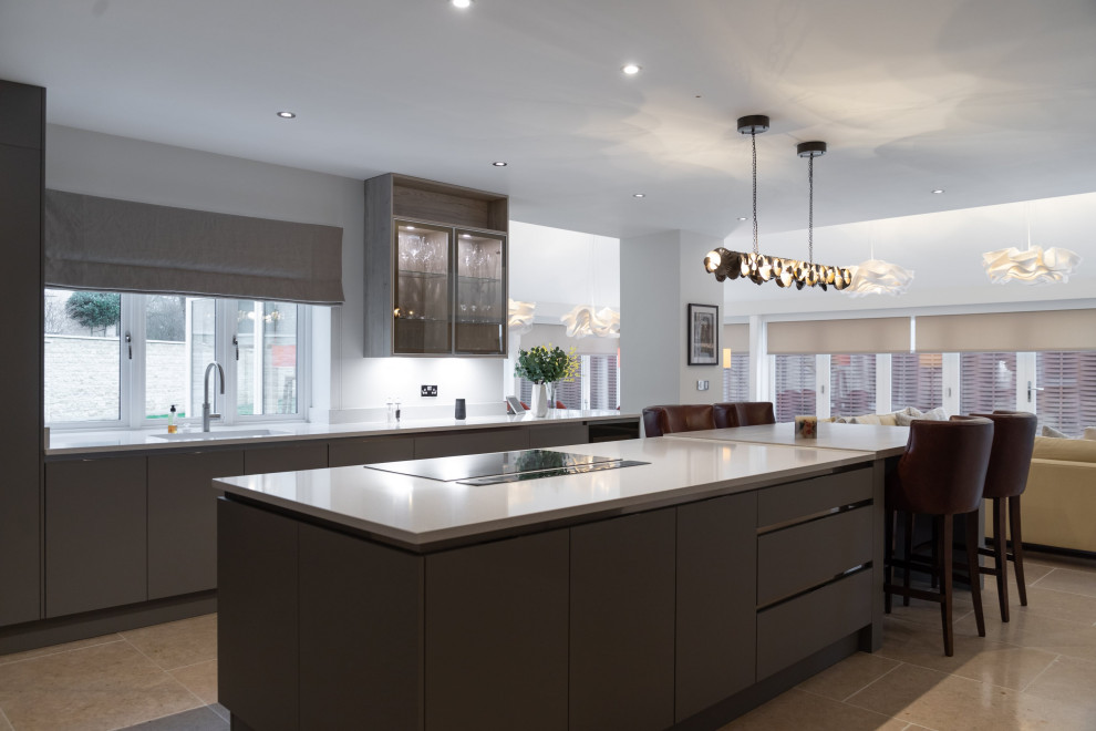 This is an example of a large contemporary grey and white open plan kitchen in London with a submerged sink, flat-panel cabinets, grey cabinets, quartz worktops, white splashback, glass sheet splashback, stainless steel appliances, limestone flooring, an island, beige floors, white worktops, a vaulted ceiling and feature lighting.