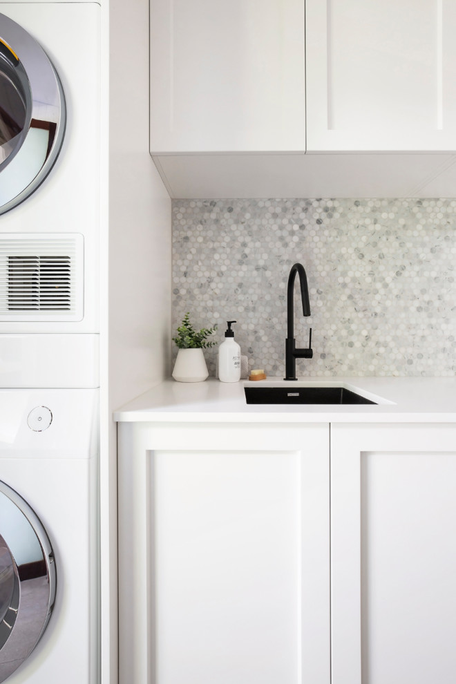 Photo of a mid-sized beach style galley dedicated laundry room in Sydney with an undermount sink, shaker cabinets, white cabinets, quartz benchtops, grey splashback, mosaic tile splashback, white walls, porcelain floors, a stacked washer and dryer, grey floor, white benchtop, coffered and panelled walls.