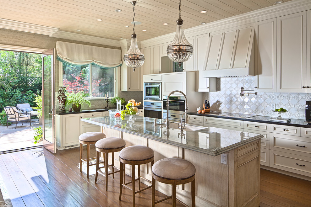 Example of a classic l-shaped medium tone wood floor and brown floor kitchen design in Los Angeles with an undermount sink, recessed-panel cabinets, beige cabinets, white backsplash, stainless steel appliances, an island and black countertops