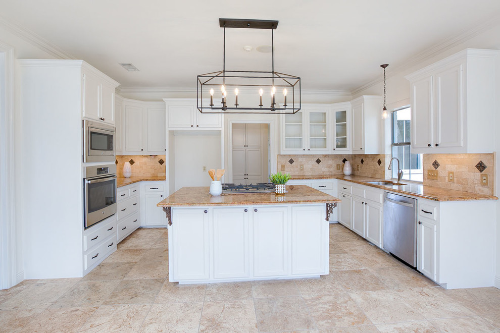 This is an example of a mid-sized traditional u-shaped open plan kitchen in New Orleans with an undermount sink, raised-panel cabinets, white cabinets, granite benchtops, beige splashback, travertine splashback, stainless steel appliances, travertine floors, with island, beige floor and beige benchtop.