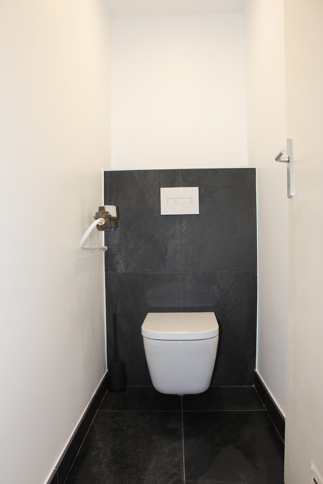 This is an example of a contemporary powder room in Grenoble with a wall-mount toilet, black tile, ceramic tile, white walls, ceramic floors, tile benchtops, black floor and black benchtops.