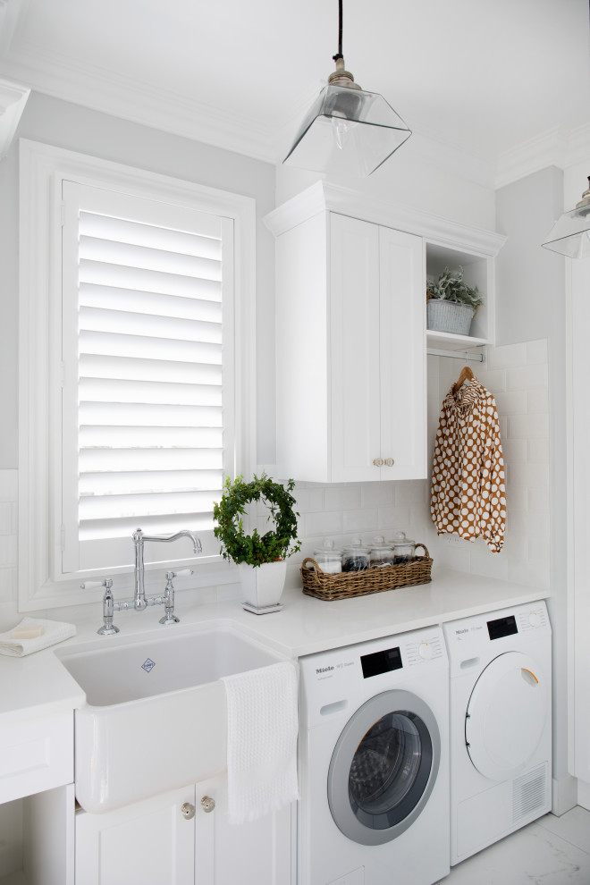 Mid-sized transitional single-wall dedicated laundry room in Brisbane with a farmhouse sink, shaker cabinets, white cabinets, white splashback, subway tile splashback, grey walls, a side-by-side washer and dryer, grey floor and white benchtop.