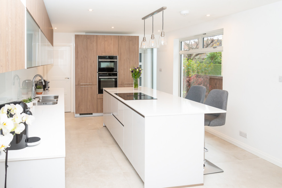 Design ideas for a mid-sized contemporary galley eat-in kitchen in Berkshire with a triple-bowl sink, flat-panel cabinets, grey cabinets, quartzite benchtops, white splashback, glass sheet splashback, black appliances, porcelain floors, with island and white benchtop.