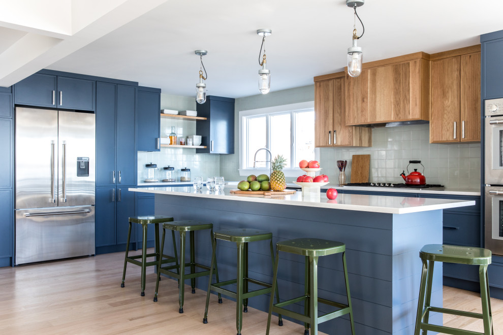 Design ideas for a large transitional l-shaped eat-in kitchen in Portland Maine with a farmhouse sink, blue cabinets, grey splashback, ceramic splashback, stainless steel appliances, light hardwood floors, with island, brown floor and white benchtop.