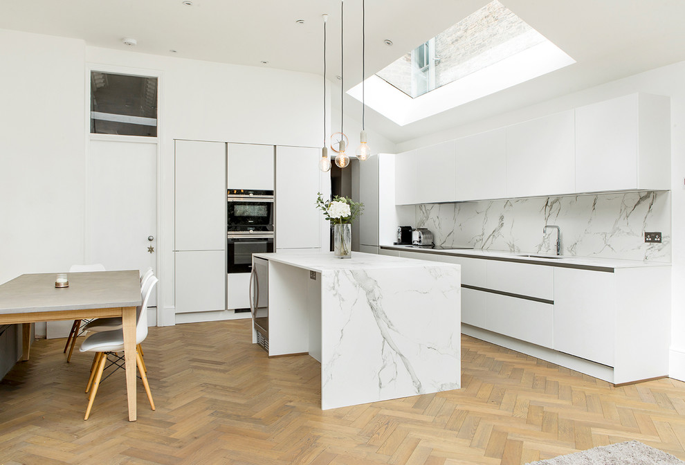 Inspiration for a mid-sized contemporary l-shaped eat-in kitchen in London with flat-panel cabinets, white cabinets, marble benchtops, white splashback, marble splashback, black appliances, light hardwood floors, with island, an undermount sink, beige floor and white benchtop.