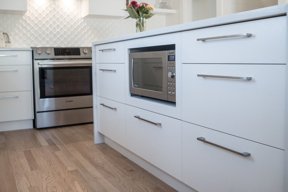 This is an example of a mid-sized transitional l-shaped eat-in kitchen in Vancouver with an undermount sink, flat-panel cabinets, white cabinets, quartz benchtops, white splashback, ceramic splashback, stainless steel appliances, light hardwood floors and with island.