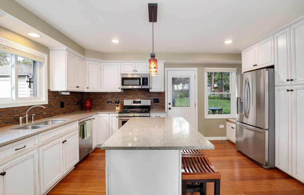 Photo of a mid-sized transitional l-shaped eat-in kitchen in Chicago with an undermount sink, raised-panel cabinets, white cabinets, quartz benchtops, multi-coloured splashback, glass tile splashback, stainless steel appliances, light hardwood floors, with island, grey benchtop, brown floor and wallpaper.