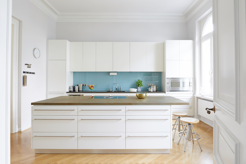 Inspiration for a large transitional galley separate kitchen in Bonn with flat-panel cabinets, white cabinets, glass sheet splashback, stainless steel appliances, light hardwood floors, brown benchtop, blue splashback, with island and beige floor.
