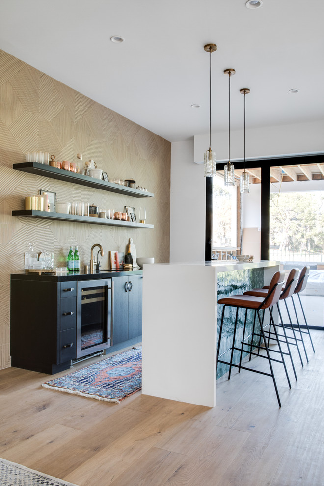 This is an example of a contemporary single-wall wet bar in Orange County with flat-panel cabinets, black cabinets, beige splashback, light hardwood floors, beige floor and black benchtop.