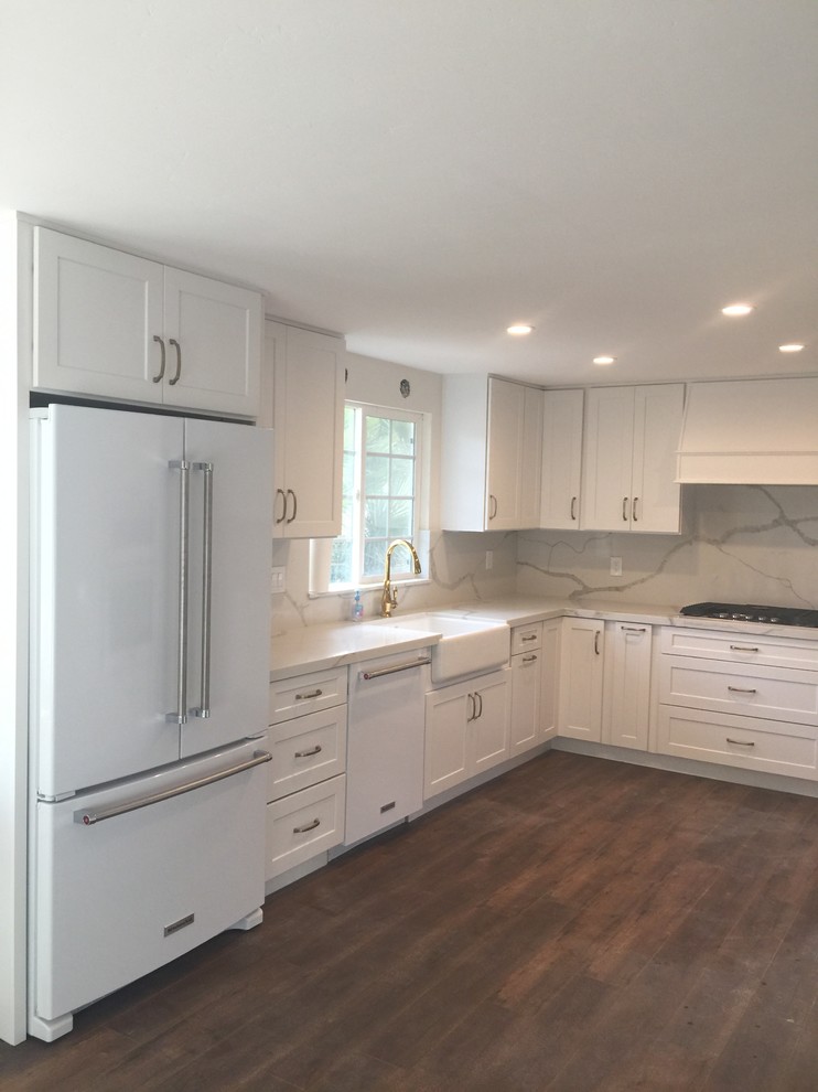 Design ideas for a large contemporary u-shaped separate kitchen in San Diego with a farmhouse sink, shaker cabinets, white cabinets, quartzite benchtops, white splashback, stone slab splashback, white appliances, medium hardwood floors, no island, brown floor and white benchtop.