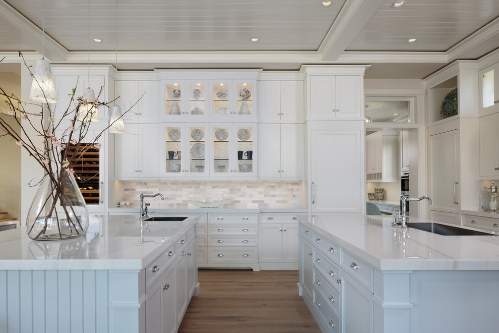 This is an example of a large tropical l-shaped eat-in kitchen in Miami with an undermount sink, raised-panel cabinets, white cabinets, marble benchtops, multi-coloured splashback, porcelain splashback, white appliances, light hardwood floors, multiple islands, brown floor and white benchtop.