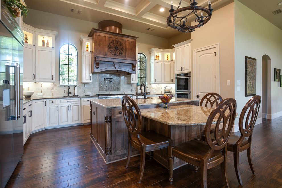Photo of an expansive traditional l-shaped open plan kitchen in Dallas with a farmhouse sink, louvered cabinets, white cabinets, granite benchtops, white splashback, cement tile splashback, stainless steel appliances, dark hardwood floors and multiple islands.