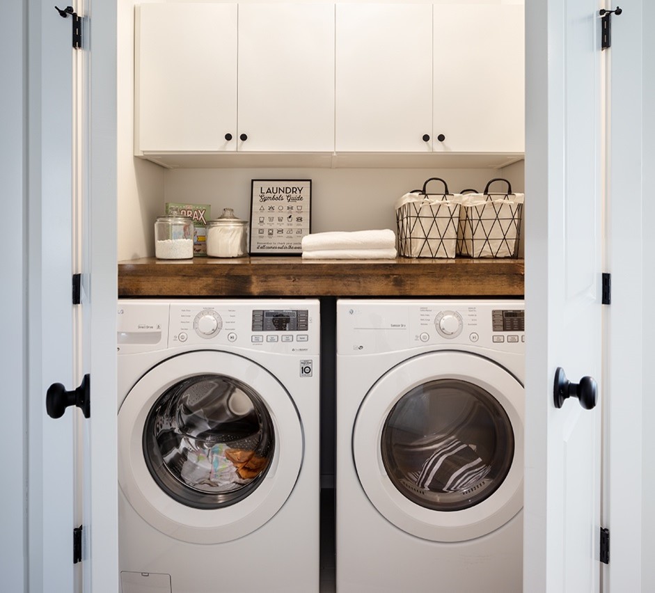 This is an example of a small country laundry cupboard in Vancouver with flat-panel cabinets, white cabinets, wood benchtops, white walls, porcelain floors, a side-by-side washer and dryer and grey floor.