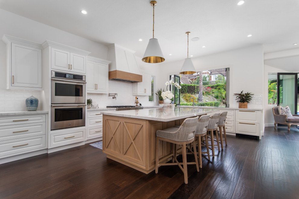 Large transitional l-shaped open plan kitchen in Orange County with raised-panel cabinets, white cabinets, stainless steel appliances, dark hardwood floors, with island, brown floor, white benchtop, an undermount sink, quartzite benchtops, white splashback and ceramic splashback.