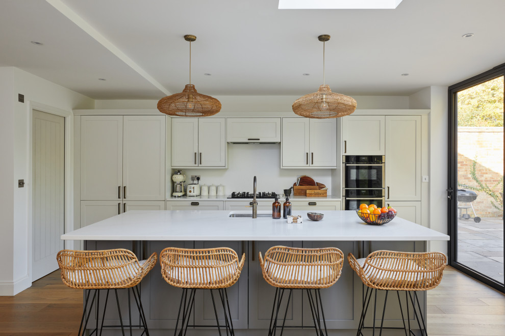 This is an example of a modern single-wall kitchen/diner in Surrey with a built-in sink, shaker cabinets, white cabinets, white splashback, medium hardwood flooring, an island, brown floors and white worktops.