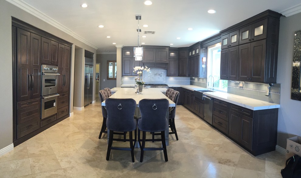 Enclosed kitchen - mid-sized transitional l-shaped travertine floor and beige floor enclosed kitchen idea in Orange County with a farmhouse sink, beaded inset cabinets, brown cabinets, marble countertops, gray backsplash, glass tile backsplash, stainless steel appliances and an island