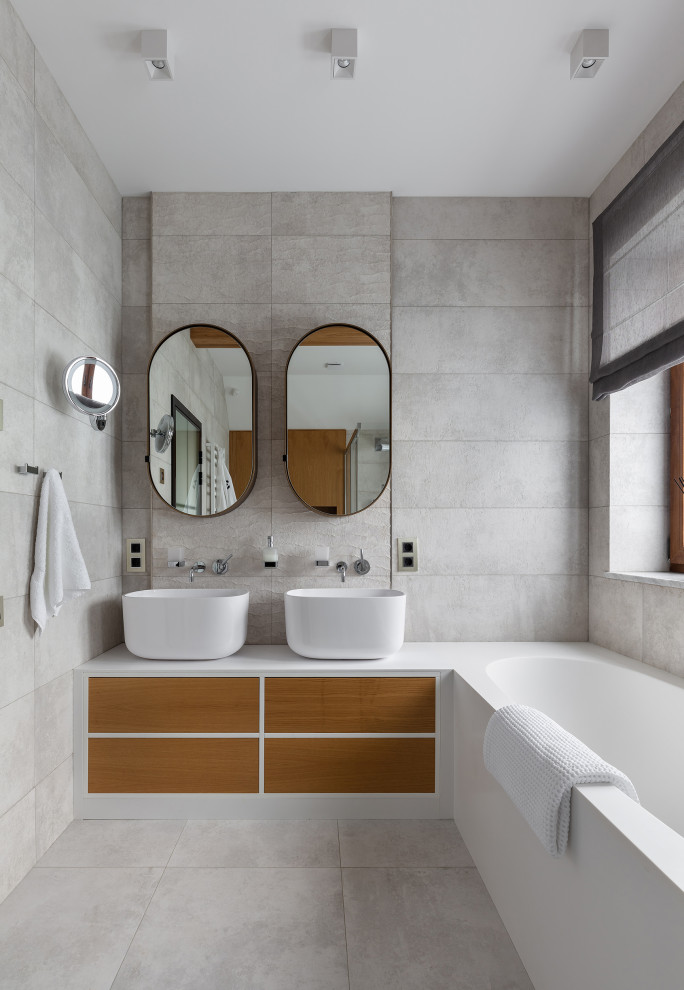 Идея дизайна: большая ванная комната в белых тонах с отделкой деревом в современном стиле с плоскими фасадами, светлыми деревянными фасадами, полновстраиваемой ванной, открытым душем, инсталляцией, серой плиткой, керамогранитной плиткой, серыми стенами, полом из керамогранита, душевой кабиной, настольной раковиной, столешницей из искусственного камня, серым полом, душем с раздвижными дверями, белой столешницей, окном, тумбой под две раковины, напольной тумбой и балками на потолке