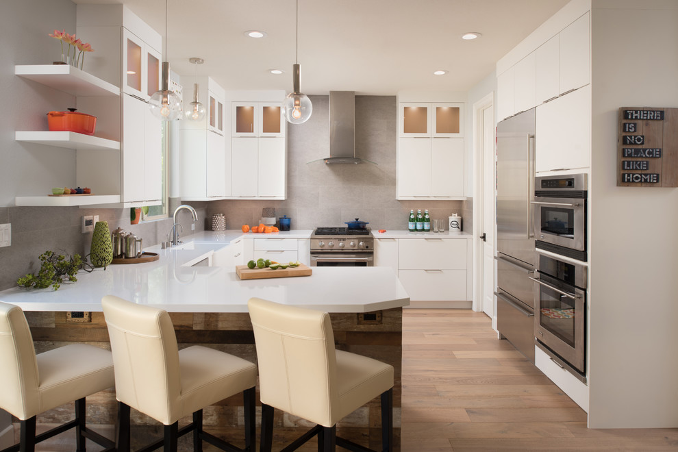 Photo of a mid-sized contemporary u-shaped open plan kitchen in San Diego with a farmhouse sink, flat-panel cabinets, white cabinets, quartz benchtops, grey splashback, porcelain splashback, stainless steel appliances and light hardwood floors.