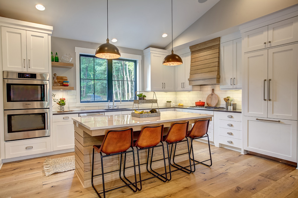 Photo of a country l-shaped kitchen in Grand Rapids with an undermount sink, shaker cabinets, white cabinets, white splashback, subway tile splashback, panelled appliances, medium hardwood floors, with island, brown floor and grey benchtop.
