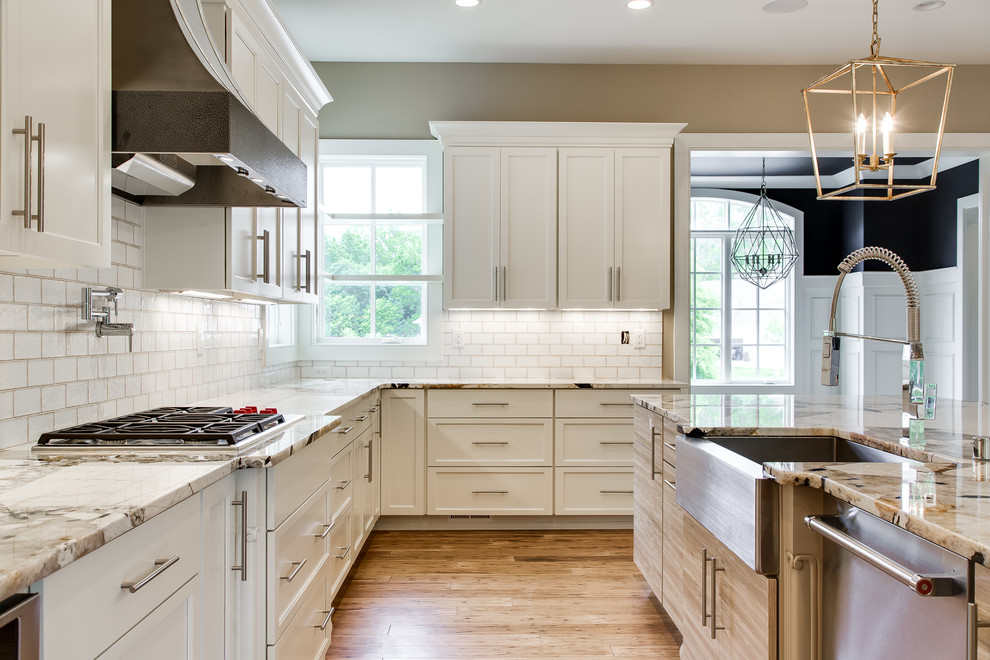 Design ideas for a mid-sized transitional l-shaped eat-in kitchen in Nashville with a farmhouse sink, recessed-panel cabinets, white cabinets, granite benchtops, white splashback, subway tile splashback, light hardwood floors, with island, brown floor and multi-coloured benchtop.