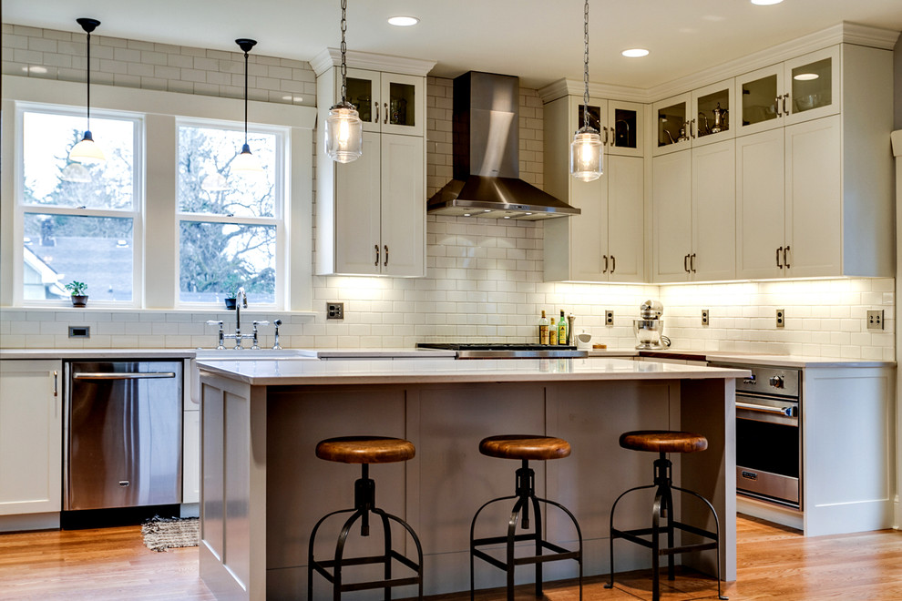 Mid-sized industrial l-shaped eat-in kitchen in Portland with a farmhouse sink, shaker cabinets, white cabinets, quartz benchtops, white splashback, ceramic splashback, stainless steel appliances, medium hardwood floors, with island and brown floor.
