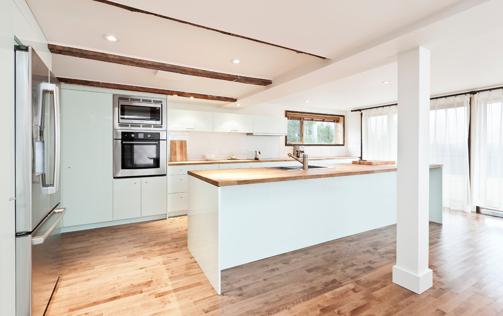 Mid-sized contemporary galley kitchen in Montreal with stainless steel appliances, a double-bowl sink, flat-panel cabinets, white cabinets, white splashback, subway tile splashback, wood benchtops, medium hardwood floors, with island and brown floor.
