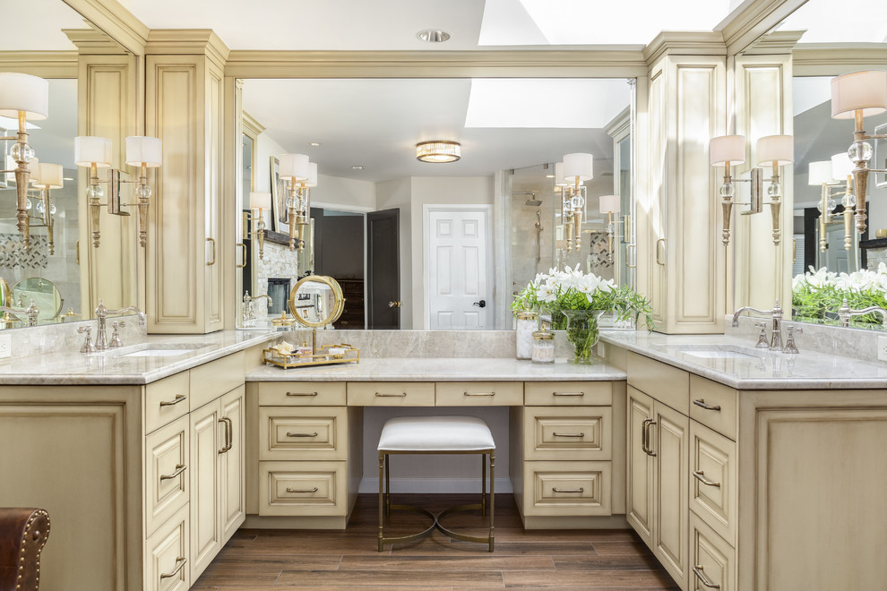 This is an example of a large transitional master bathroom in Seattle with raised-panel cabinets, distressed cabinets, a double shower, a one-piece toilet, beige tile, marble, beige walls, ceramic floors, an undermount sink, quartzite benchtops, brown floor, a hinged shower door and beige benchtops.