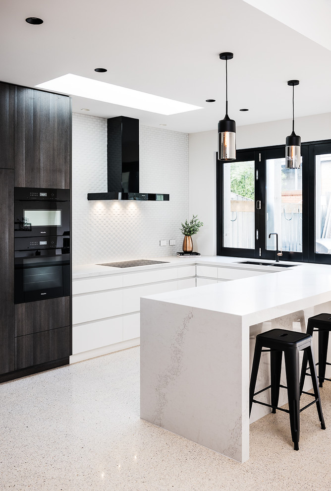 Photo of a mid-sized contemporary u-shaped eat-in kitchen in Melbourne with an undermount sink, flat-panel cabinets, white cabinets, quartz benchtops, white splashback, ceramic splashback, black appliances, concrete floors, a peninsula, beige floor and white benchtop.