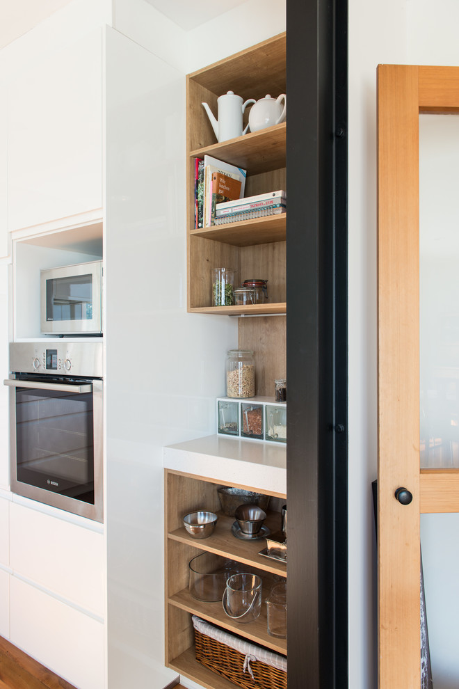 This is an example of a mid-sized scandinavian u-shaped kitchen pantry in Christchurch with an undermount sink, flat-panel cabinets, white cabinets, solid surface benchtops, glass sheet splashback, white appliances, medium hardwood floors, a peninsula and brown floor.