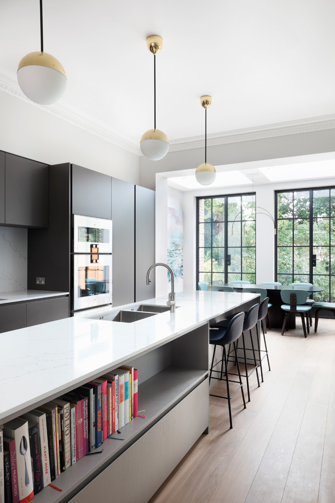 Photo of a contemporary single-wall eat-in kitchen in Buckinghamshire with flat-panel cabinets, black cabinets, quartzite benchtops, white splashback, with island, white benchtop, brown floor and medium hardwood floors.