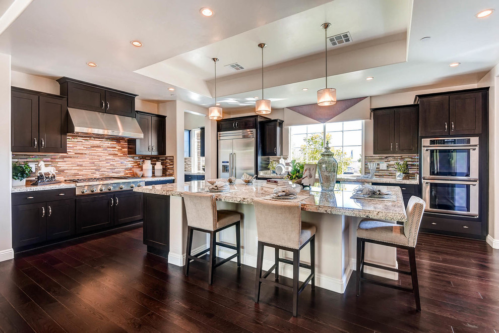 This is an example of a large transitional l-shaped open plan kitchen in Las Vegas with shaker cabinets, dark wood cabinets, granite benchtops, metallic splashback, glass tile splashback, stainless steel appliances, dark hardwood floors and with island.