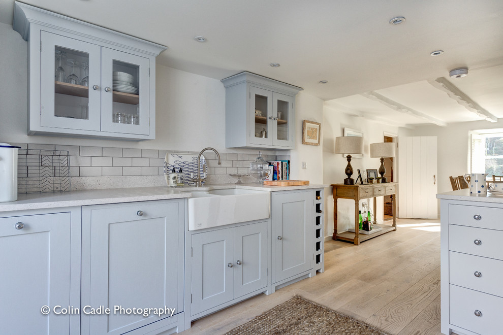 Photo of a small beach style u-shaped eat-in kitchen in Devon with a farmhouse sink, recessed-panel cabinets, distressed cabinets, marble benchtops, white splashback, ceramic splashback and light hardwood floors.