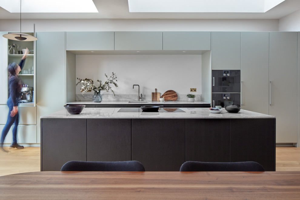 Photo of a large contemporary galley kitchen/diner in London with a submerged sink, flat-panel cabinets, marble worktops, marble splashback, an island, grey cabinets, black appliances, medium hardwood flooring, brown floors and white worktops.