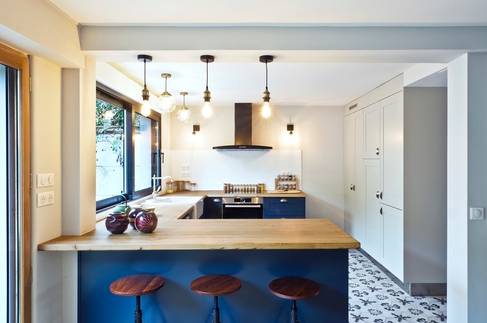 Mid-sized transitional u-shaped kitchen in Paris with wood benchtops, white splashback, stainless steel appliances, cement tiles, a farmhouse sink, shaker cabinets, blue cabinets, a peninsula, multi-coloured floor and beige benchtop.