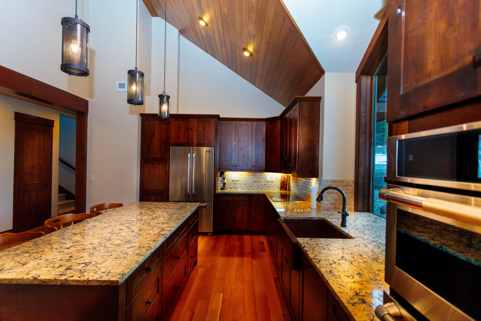 This is an example of a mid-sized country l-shaped open plan kitchen in Denver with a farmhouse sink, shaker cabinets, dark wood cabinets, granite benchtops, beige splashback, subway tile splashback, stainless steel appliances, medium hardwood floors, with island, brown floor and multi-coloured benchtop.