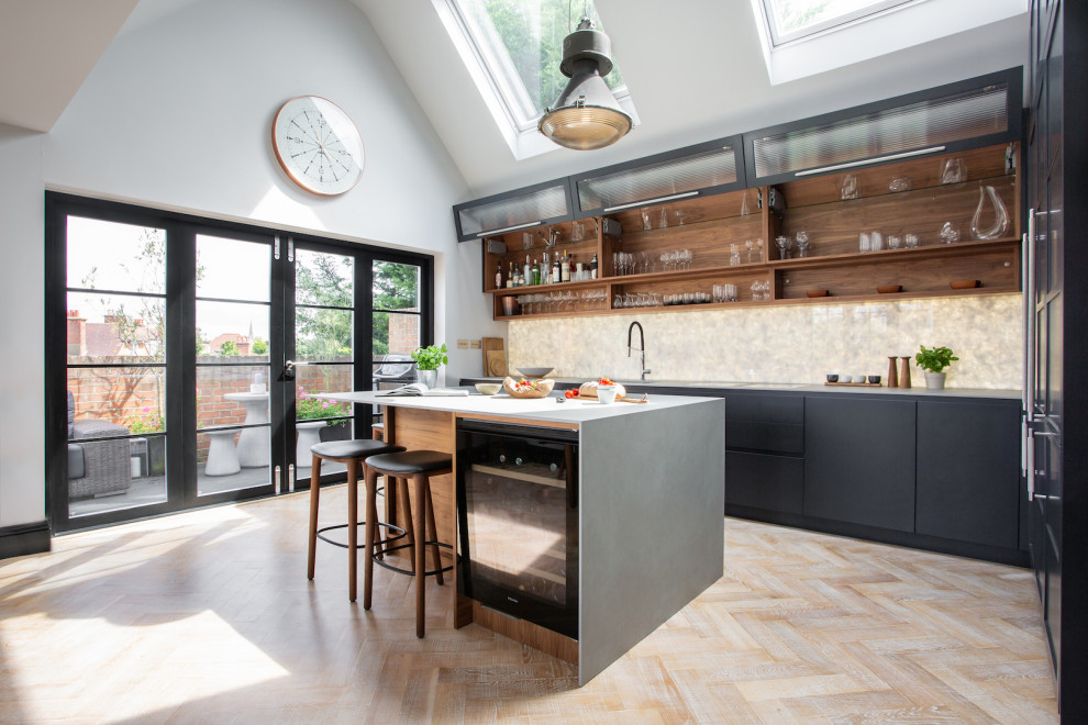 Small transitional open plan kitchen in London with raised-panel cabinets, solid surface benchtops, white splashback, marble splashback, black appliances, light hardwood floors, with island and grey benchtop.