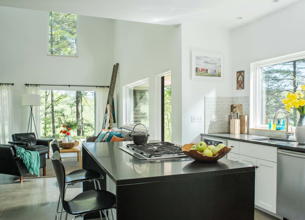 Photo of a small contemporary single-wall open plan kitchen in Burlington with a single-bowl sink, shaker cabinets, white cabinets, solid surface benchtops, grey splashback, subway tile splashback, stainless steel appliances, concrete floors, with island, grey floor and black benchtop.