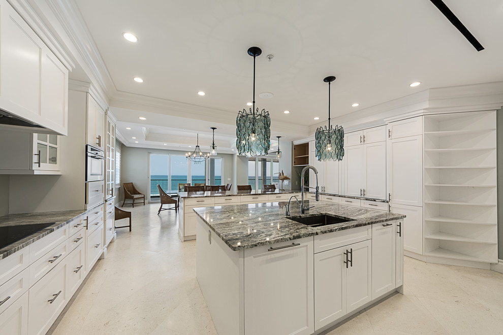 Design ideas for an expansive traditional u-shaped open plan kitchen in Miami with an undermount sink, recessed-panel cabinets, white cabinets, granite benchtops, grey splashback, cement tile splashback, stainless steel appliances, limestone floors, with island, beige floor and grey benchtop.