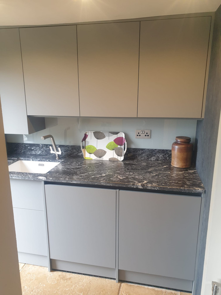 Inspiration for a mid-sized contemporary l-shaped laundry room in West Midlands with flat-panel cabinets, granite benchtops, black splashback, engineered quartz splashback, black benchtop, a drop-in sink, black cabinets, travertine floors, beige floor and vaulted.