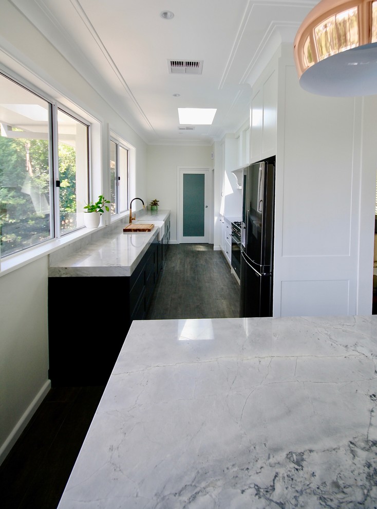 This is an example of a large transitional galley eat-in kitchen in Sydney with a farmhouse sink, shaker cabinets, black cabinets, quartzite benchtops, white splashback, ceramic splashback, stainless steel appliances, medium hardwood floors, with island, grey floor and grey benchtop.