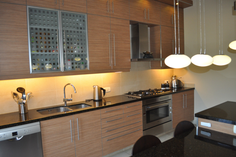 Mid-sized minimalist galley ceramic tile and beige floor enclosed kitchen photo with a double-bowl sink, flat-panel cabinets, medium tone wood cabinets, granite countertops, beige backsplash, ceramic backsplash, stainless steel appliances, an island and black countertops