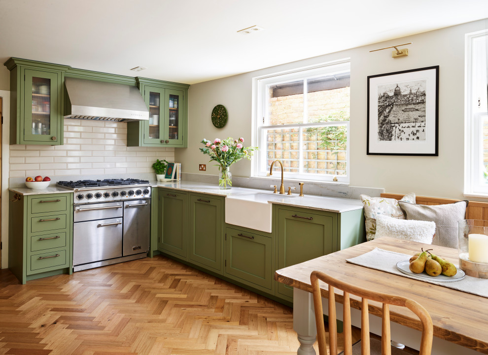 Design ideas for a classic l-shaped kitchen/diner in London with shaker cabinets, a belfast sink, green cabinets, white splashback, stainless steel appliances, medium hardwood flooring, brown floors and white worktops.