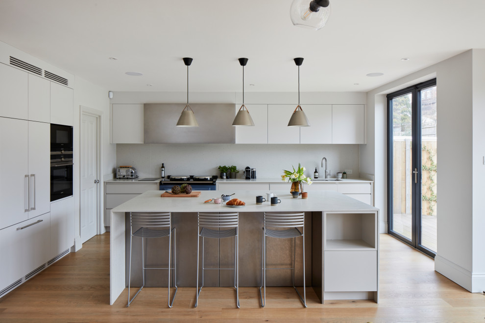 Photo of a large contemporary l-shaped kitchen/diner in London with flat-panel cabinets, white cabinets, composite countertops, white splashback, black appliances, an island, white worktops, a submerged sink, light hardwood flooring and beige floors.