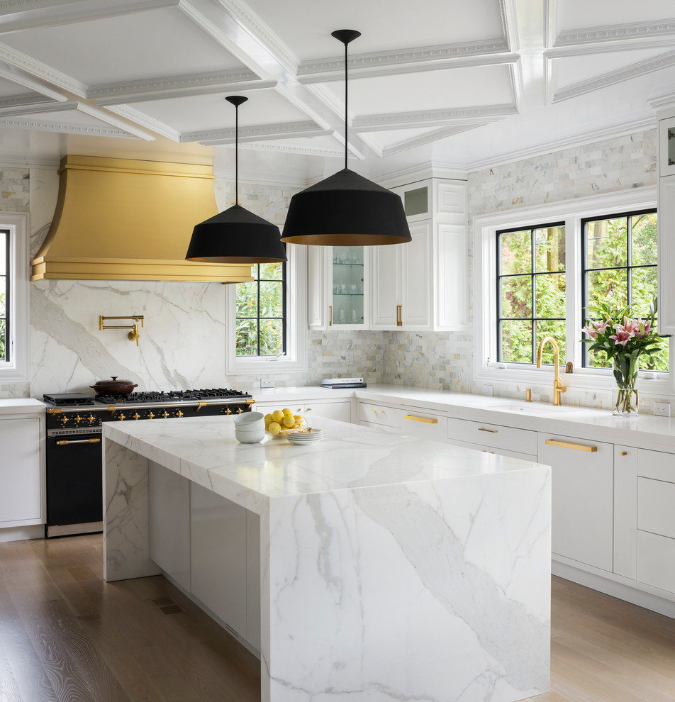 This is an example of a large transitional u-shaped kitchen in New York with a drop-in sink, flat-panel cabinets, white cabinets, marble benchtops, white splashback, marble splashback, black appliances, light hardwood floors, with island, beige floor and white benchtop.