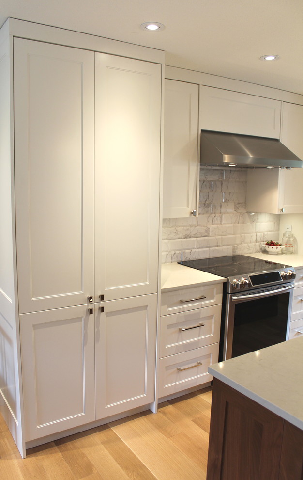 Large transitional l-shaped light wood floor eat-in kitchen photo with an undermount sink, shaker cabinets, white cabinets, quartz countertops, white backsplash, ceramic backsplash, stainless steel appliances and an island