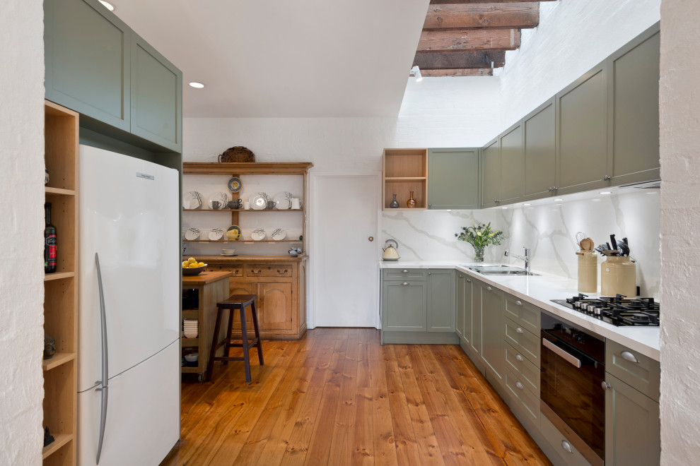 Photo of a mid-sized transitional l-shaped eat-in kitchen in Melbourne with a drop-in sink, shaker cabinets, green cabinets, quartz benchtops, white splashback, stone slab splashback, black appliances, medium hardwood floors, with island, brown floor and white benchtop.