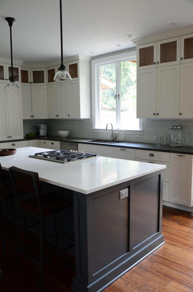 Example of a mid-sized transitional l-shaped dark wood floor eat-in kitchen design with a single-bowl sink, shaker cabinets, white cabinets, solid surface countertops, white backsplash, glass tile backsplash, stainless steel appliances and an island