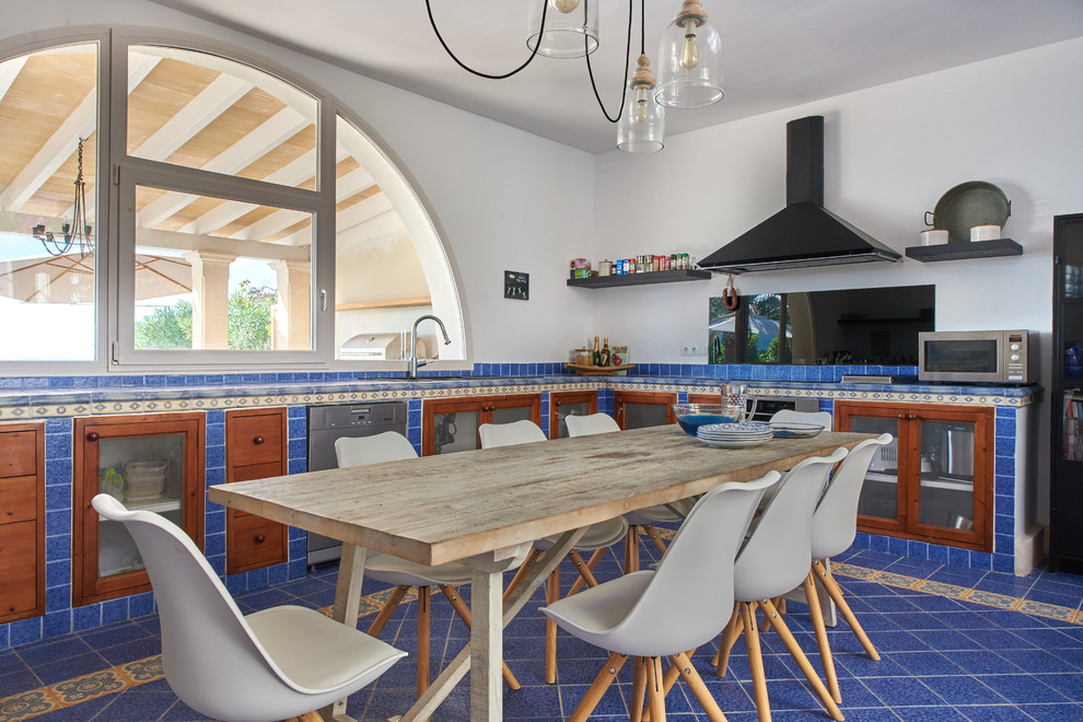 This is an example of a beach style l-shaped kitchen in Palma de Mallorca with glass-front cabinets, medium wood cabinets, an undermount sink, blue splashback, stainless steel appliances and blue floor.