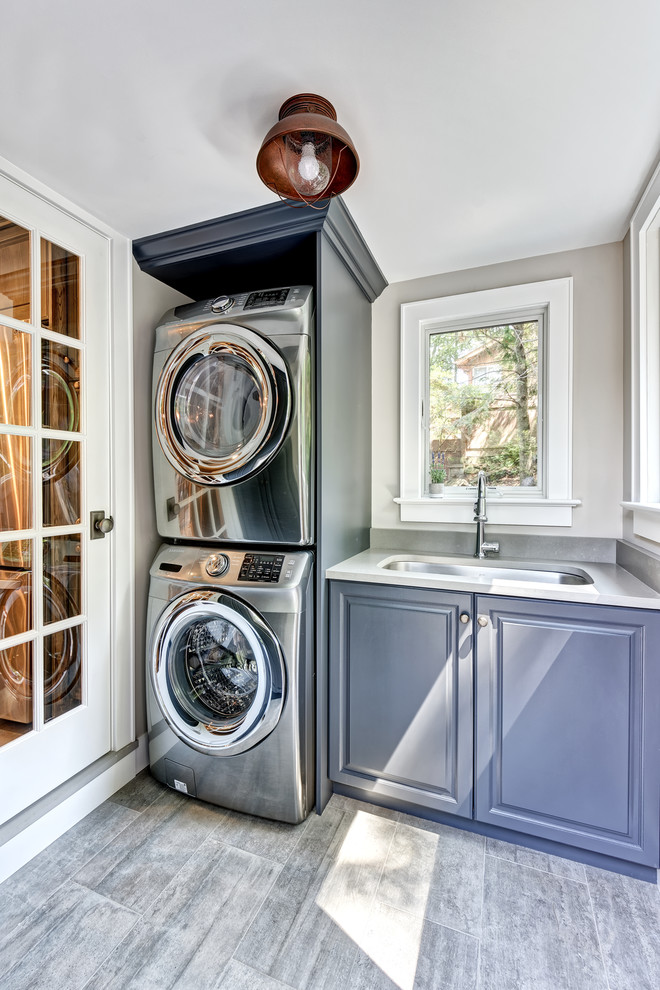 Inspiration for a small transitional single-wall utility room in New York with an undermount sink, raised-panel cabinets, grey cabinets, quartz benchtops, grey walls, porcelain floors, a stacked washer and dryer, grey floor and white benchtop.